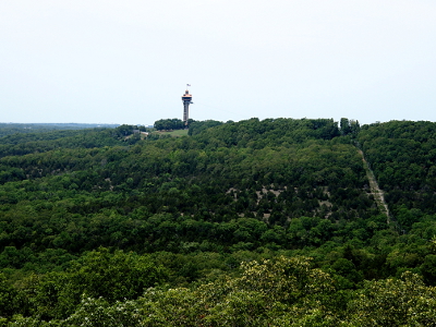 [A tall, circular tower with a flag flying atop it stands approximately five times as high as the trees around it. To the right The zipline runs from the tower to the top of the hillside then makes a 90 degree turn to go down the hillside. One can see an open area in the trees where the zipline route heads down the mountain.]